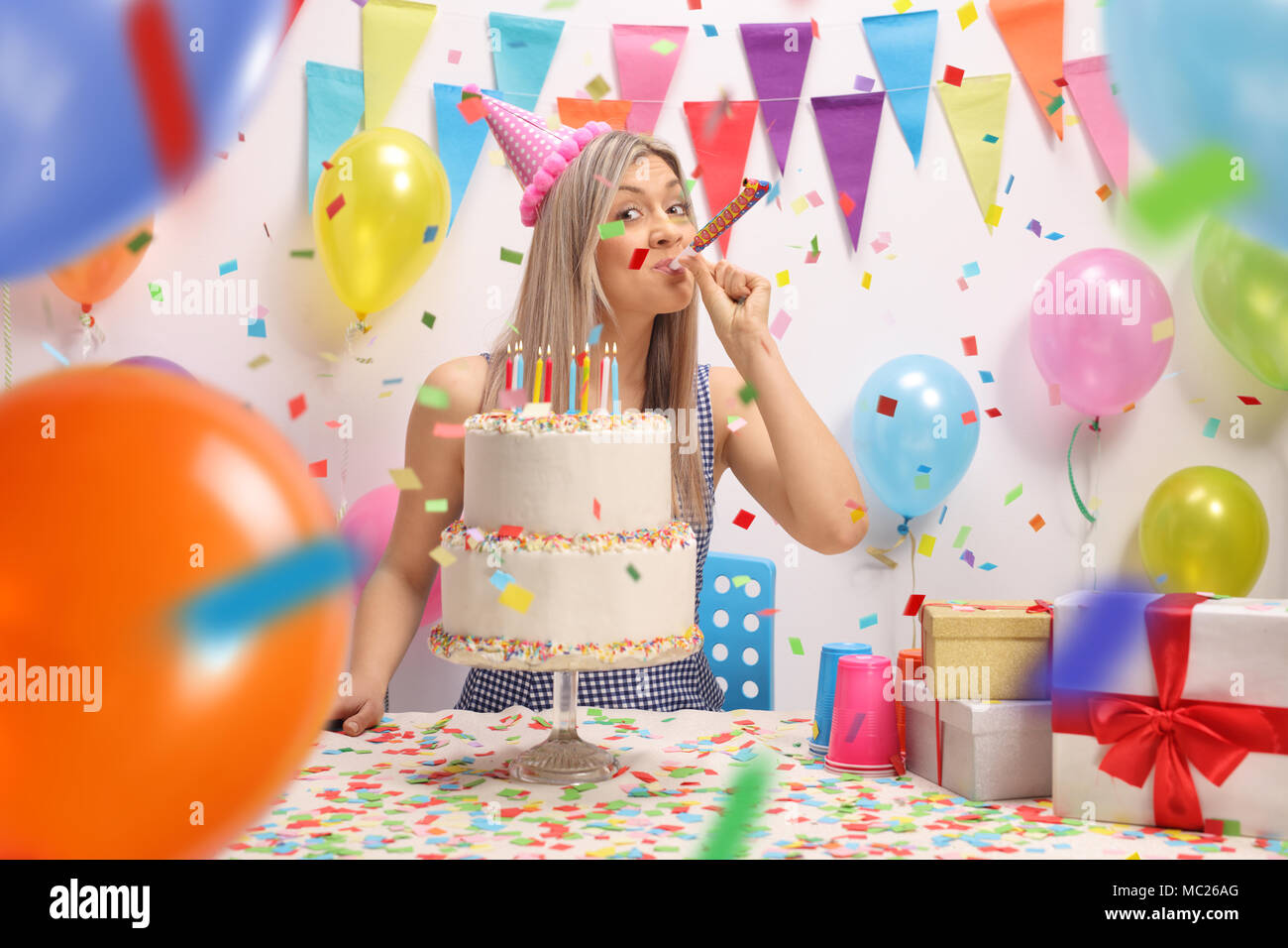 Junge Frau mit einem Geburtstagskuchen Blasen einer Partei Horn gegen eine Wand mit Ballons und Dekoration Fahnen Stockfoto