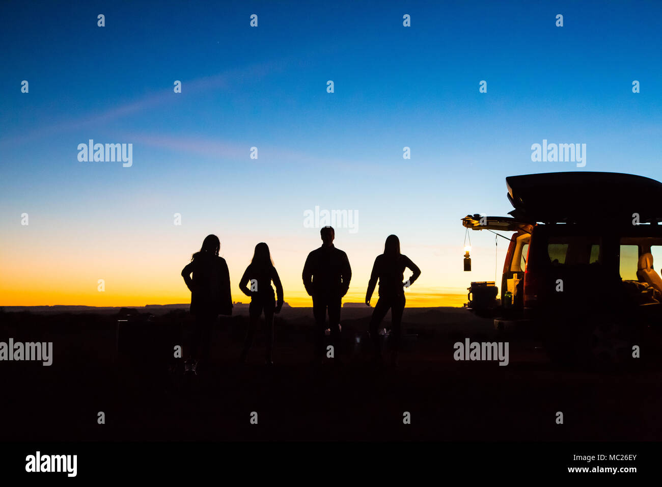 Die Silhouetten einer vierköpfigen Familie stehen hinter einem offenen Geländewagen mit einer Laterne hängend. Sie erscheinen auf dem Campingplatz. Stockfoto