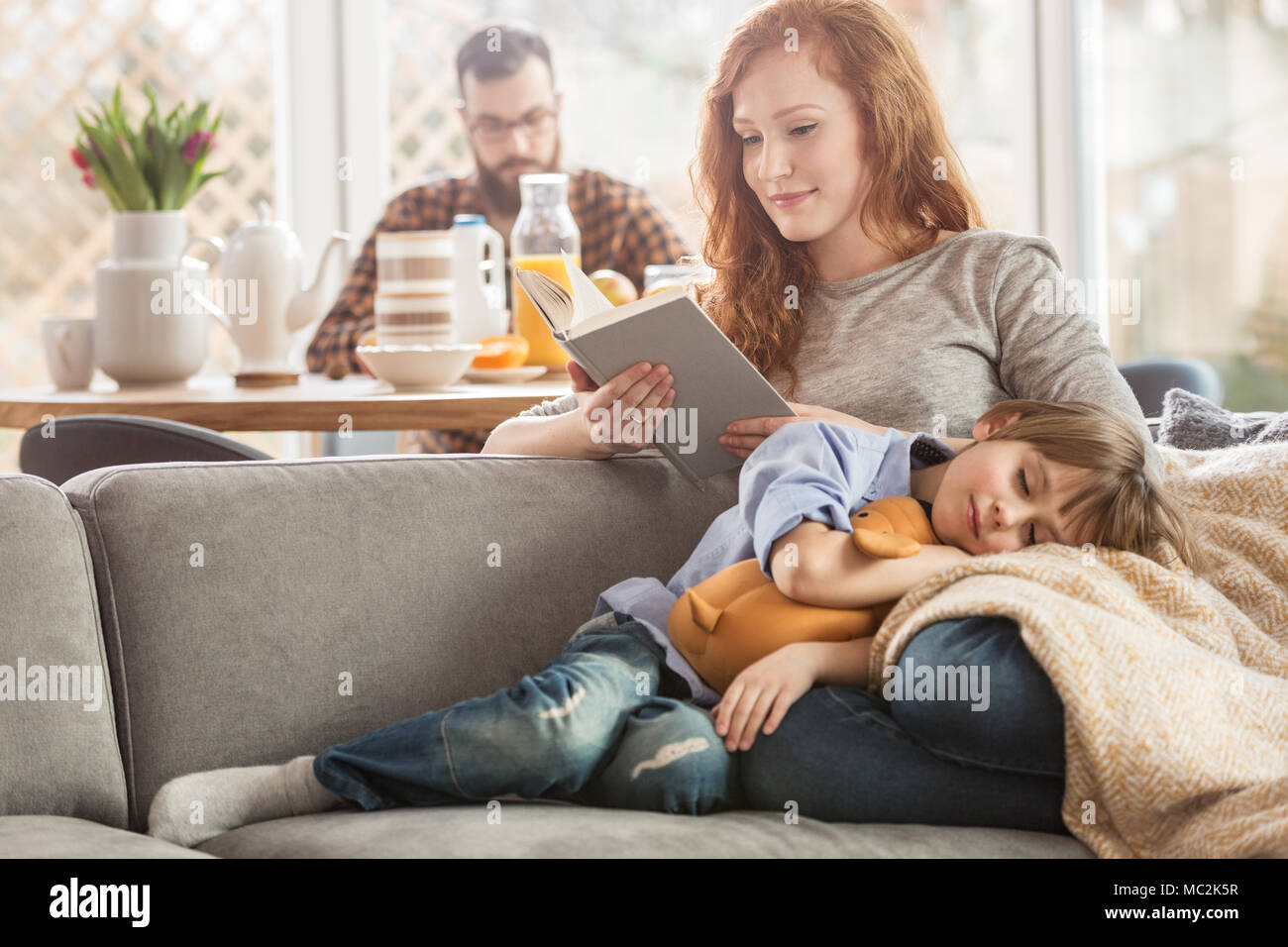 Schlafenden Sohn liegend auf Mutters Schoß, während Sie ein Buch lesen und Vater Essen im Hintergrund Stockfoto