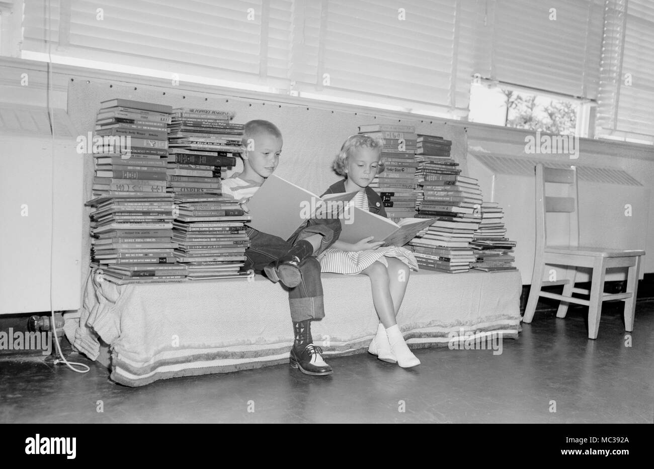 Zwei Kindern im Grundschulalter lesen Sie unter einem großen Stapel Bücher in der Schule, Ca. 1960. Stockfoto