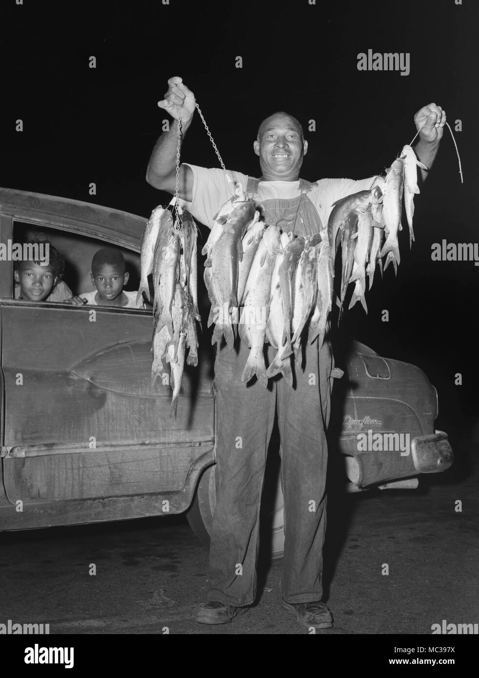 Vater zeigt stolz einen Stringer von Welsen, während sich die Kinder im Getriebe Dynaflow Buick Automobil warten, Ca. 1963. Stockfoto
