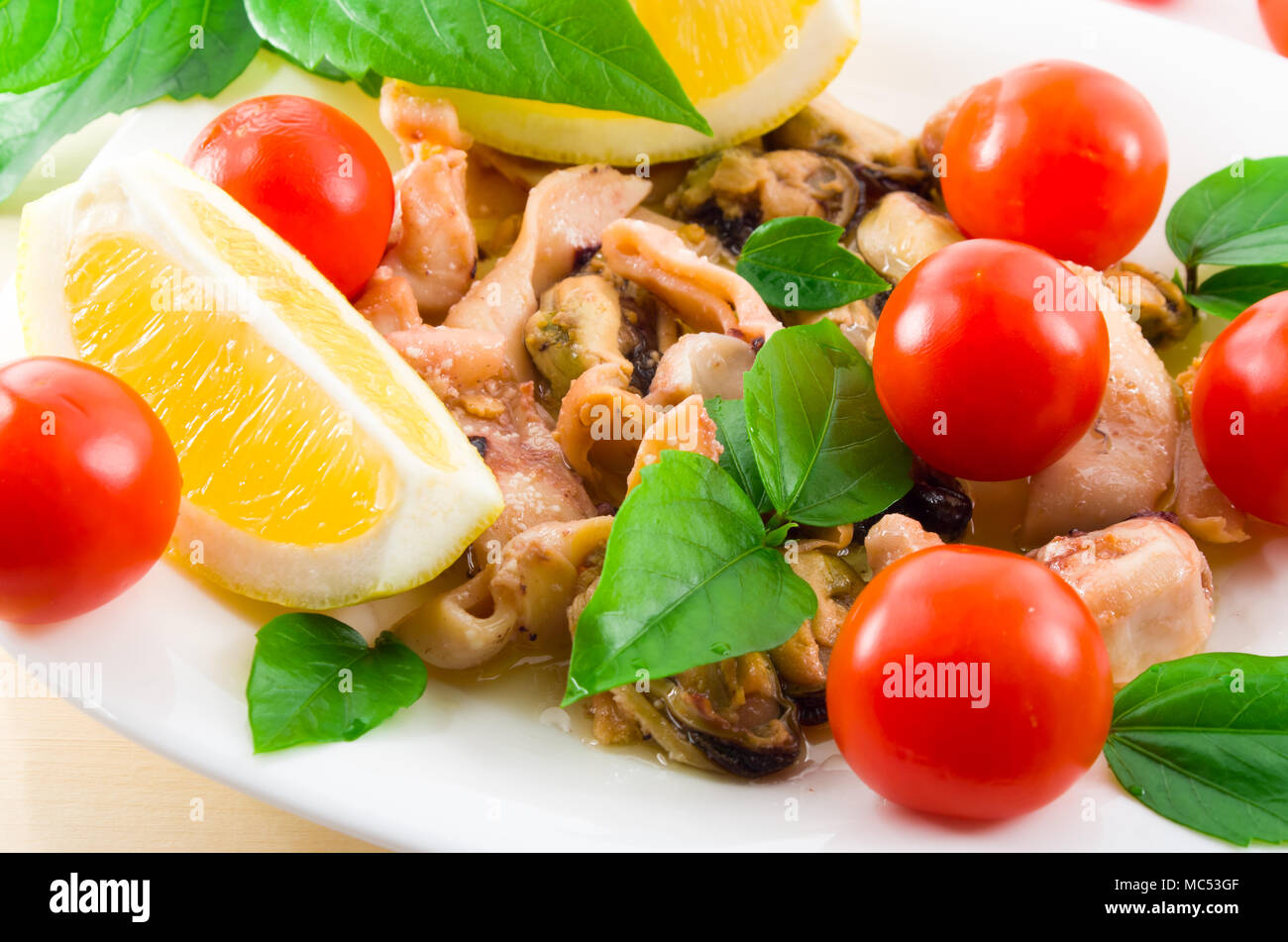 Salat von blanchierte Stücke von Meeresfrüchten auf einer weißen Platte close-up. Muscheln und Tintenfisch, dekoriert mit grünen, Cherry Tomaten und Zitronen mit Stockfoto