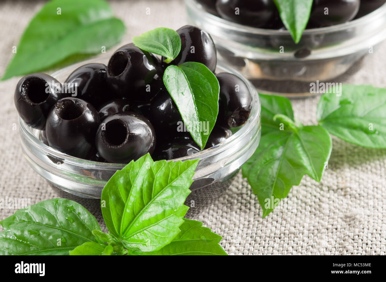Schwarze Oliven, entsteint in eine Glasschüssel auf einem Hintergrund von grauen Sackleinen und grüne Blätter marinierte Stockfoto