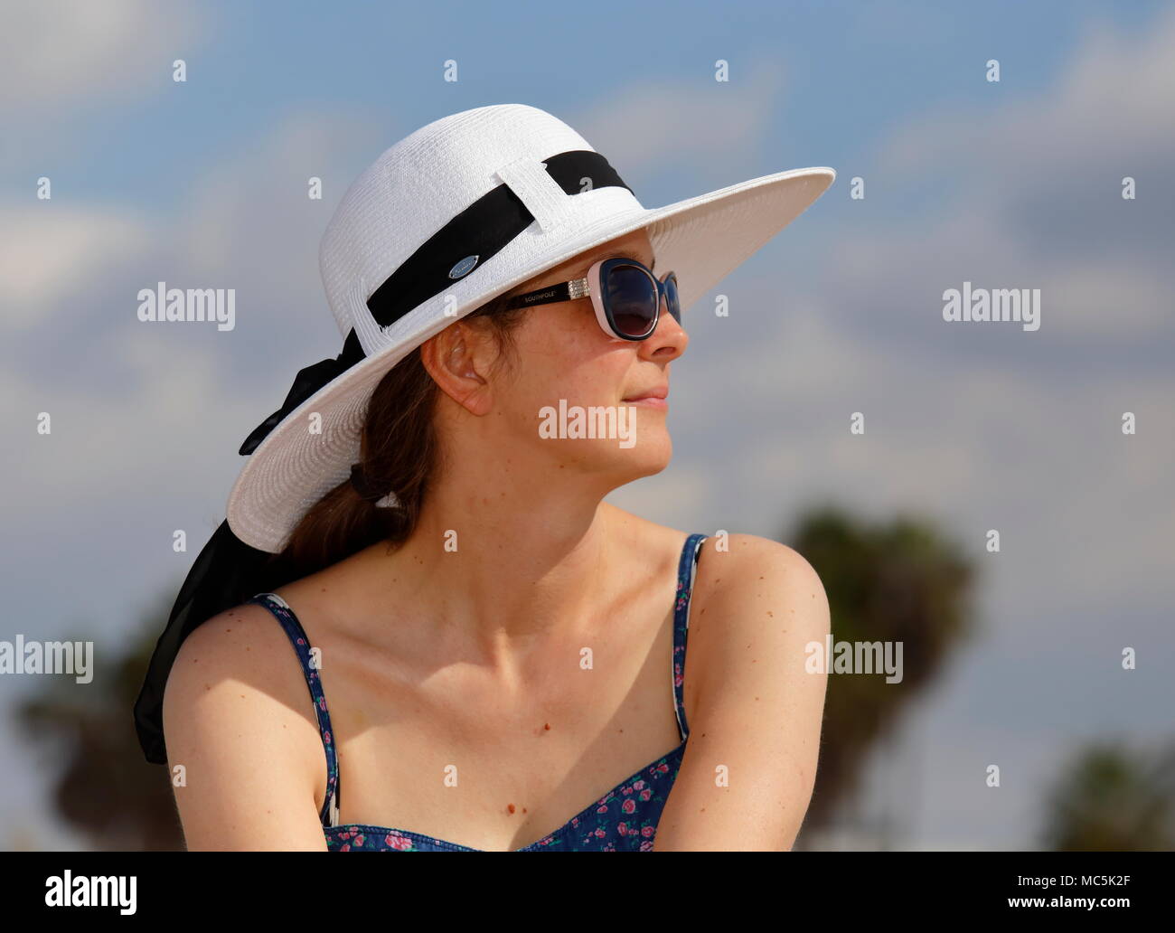 Profil anzeigen von einer jungen Frau in weißem Sonnenhut, Sonnenbrille, und blau sundress. Unscharfe Bäume, blauer Himmel und Wolken im Hintergrund Stockfoto