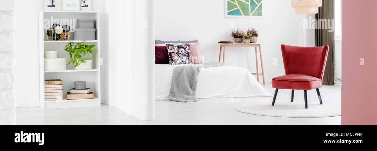 Foto von kleinen Regal mit Büchern, Pflanzen und Einrichtung und weißen Schlafzimmer Einrichtung mit burgunderroten Sessel hinter einer Wand Rohre Stockfoto