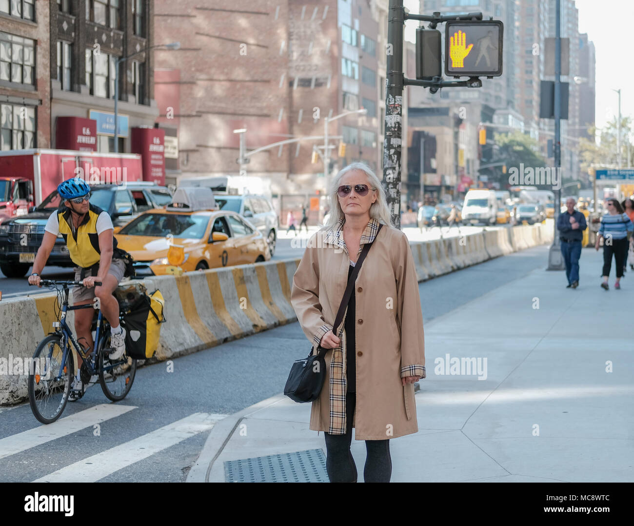 Frau mittleren Alters gesehen, eine berühmte Marke Mantel an einer Kreuzung in New York City. Der Hintergrund zeigt ein Radfahrer und ein Yellow Cab. Stockfoto