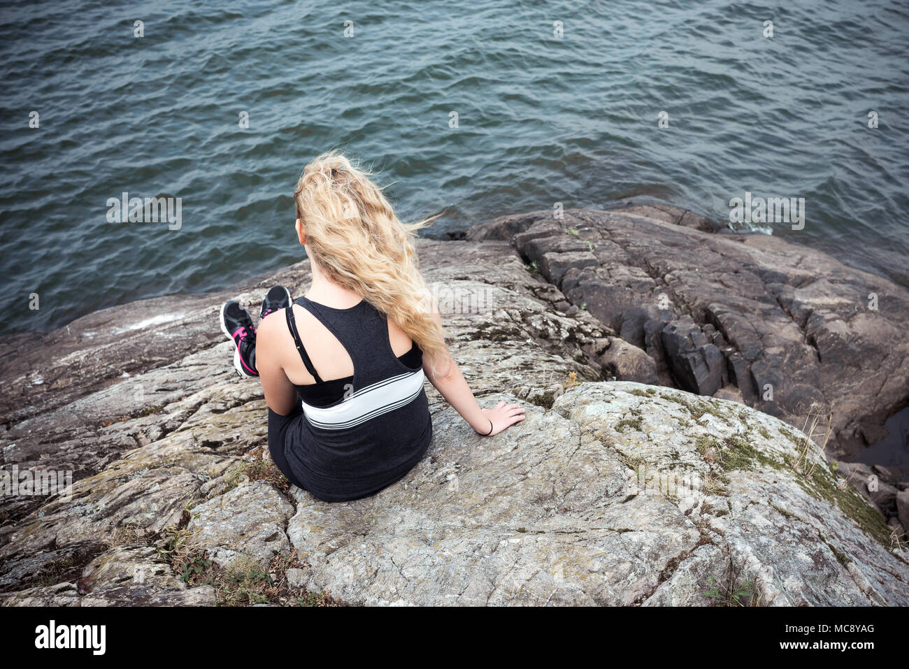 Ansicht der Rückseite eine blonde junge Frau sitzt auf einem Felsen mit Blick auf der Suche Stockfoto