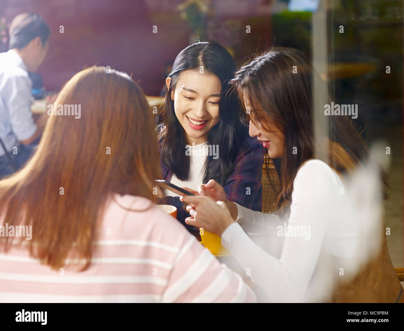 Drei glückliche wunderschöne junge asiatische Frauen am Tisch sitzen, plaudern, spielen mit Handy in ein Cafe oder Tee Haus, Schuß durch das Fenster Glas Stockfoto