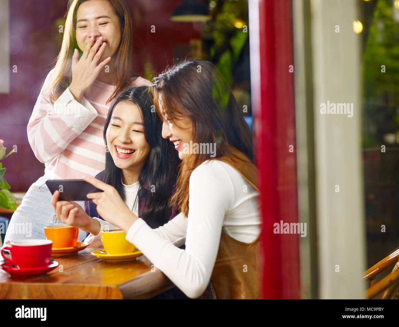 Drei glückliche wunderschöne junge asiatische Frauen am Tisch sitzen, Plaudern, Lachen, Spielen mit Handy in ein Cafe oder Tee Haus, Schuß durch wi Stockfoto