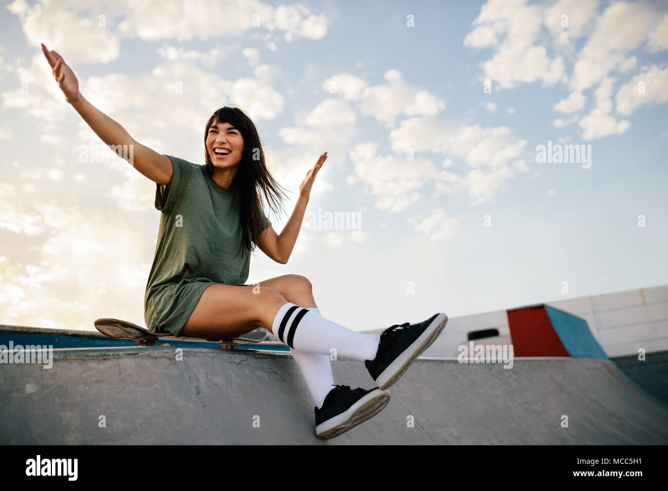 Schöne Frau auf einem Skateboard sitzen auf Rampe in Skate Park weg schauen und Lachen. Frau skateboarder genießen Sie einen Tag am Skatepark. Stockfoto
