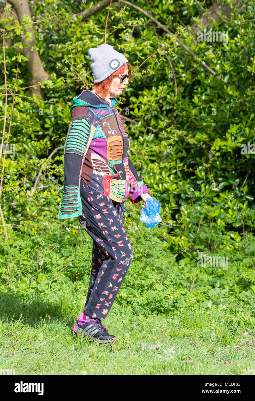 Eine hell gekleidete Mitte für ältere ältere Frau im Alter das Tragen einer Mütze zu Fuß durch den Wald an einem sonnigen Frühlingstag in England, Großbritannien. Senior trendige Kommode. Stockfoto