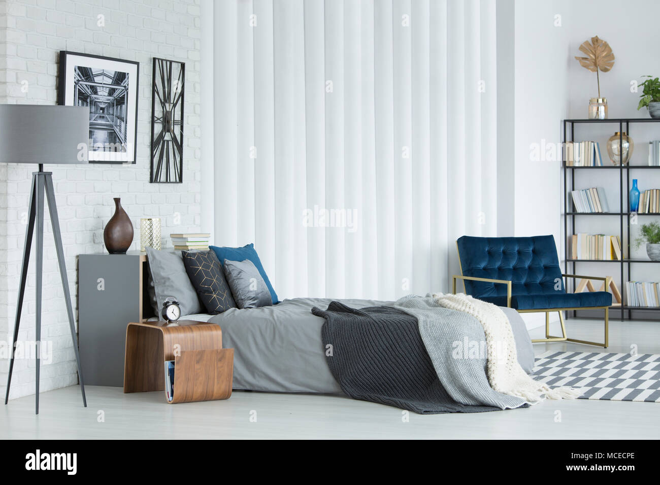 Grau Lampe neben dem Bett gegen weiße Wand mit Plakat und Wecker im Schlafzimmer Innenraum mit Sessel Stockfoto
