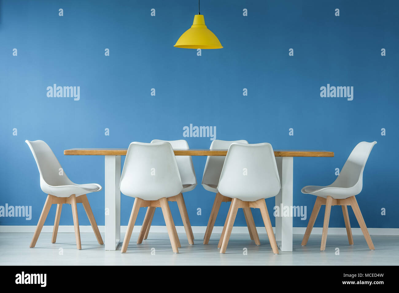 Modernes, weißes und Stühle aus Holz um einen Esstisch und gelbe Lampe vor blauem Hintergrund Wand in einem minimalistischen Interieur. Stockfoto