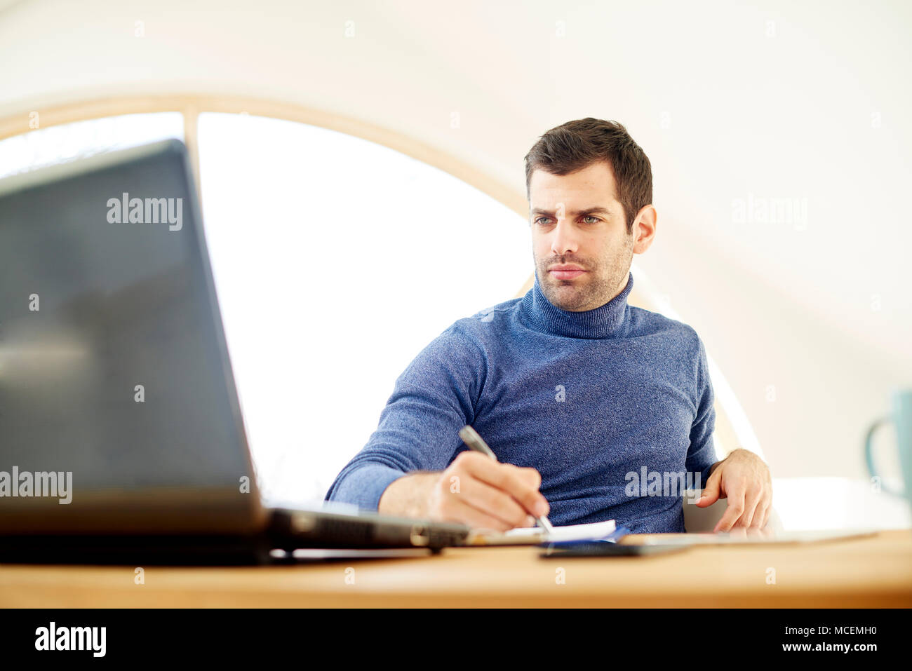 Portrait von Casual jungen Mann mit Rollkragen Pullover und Suchen thoughtul. Arbeiten am Laptop während des Schreibens mit Stift Dokumente. Home Office. Stockfoto