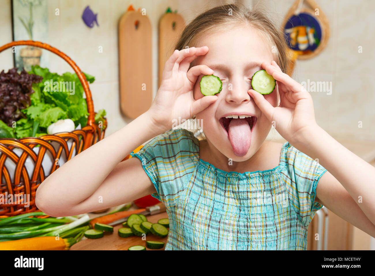 Mädchen schließt die Augen mit Gurken und viel Spaß in der Küche zu Hause. Korb mit Gemüse und frisches Obst in der Küche Interieur. Gesunde Ernährung Konzept Stockfoto