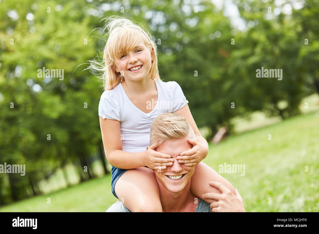 Blonde Mädchen sitzt huckepack auf Vater und hält seine Augen geschlossen. Stockfoto