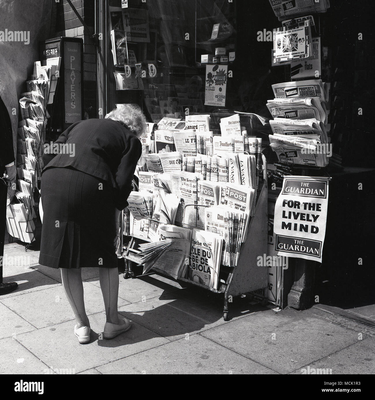 London, 1967, eine Dame beugt sich, um die Schlagzeilen der internationalen, nationalen und lokalen britischen Zeitungen wie der Kensington Post auf einem Zeitungsstand vor einem Tabakkioskiosks zu lesen. Am Stand sind Glücksmedaillons von „World Cup Willie“ zu sehen, Souvenirs für das Fußballweltcup-Turnier 1966, das in Großbritannien stattfand, mit dem Finale im Wembley Stadium. Stockfoto