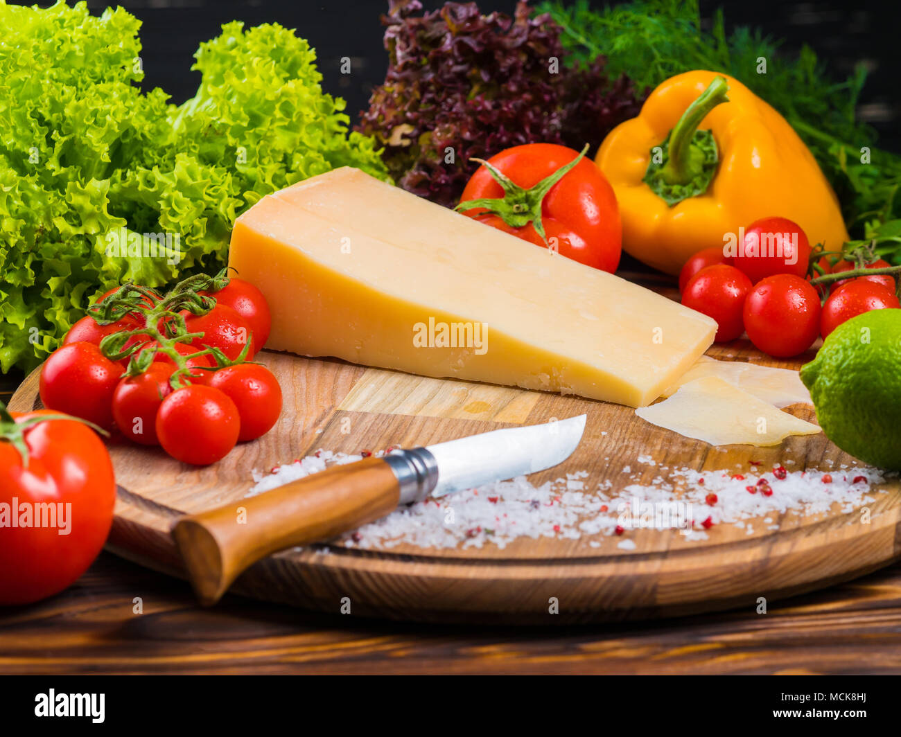 Holzbrett mit Parmesan Käse, frische Tomaten, Kopfsalat, Meersalz, Paprika und rote bulgarischen Pfeffer. Das Messer zum Schneiden von Gemüse. Noch appetitlich Leben von Gemüse Stockfoto