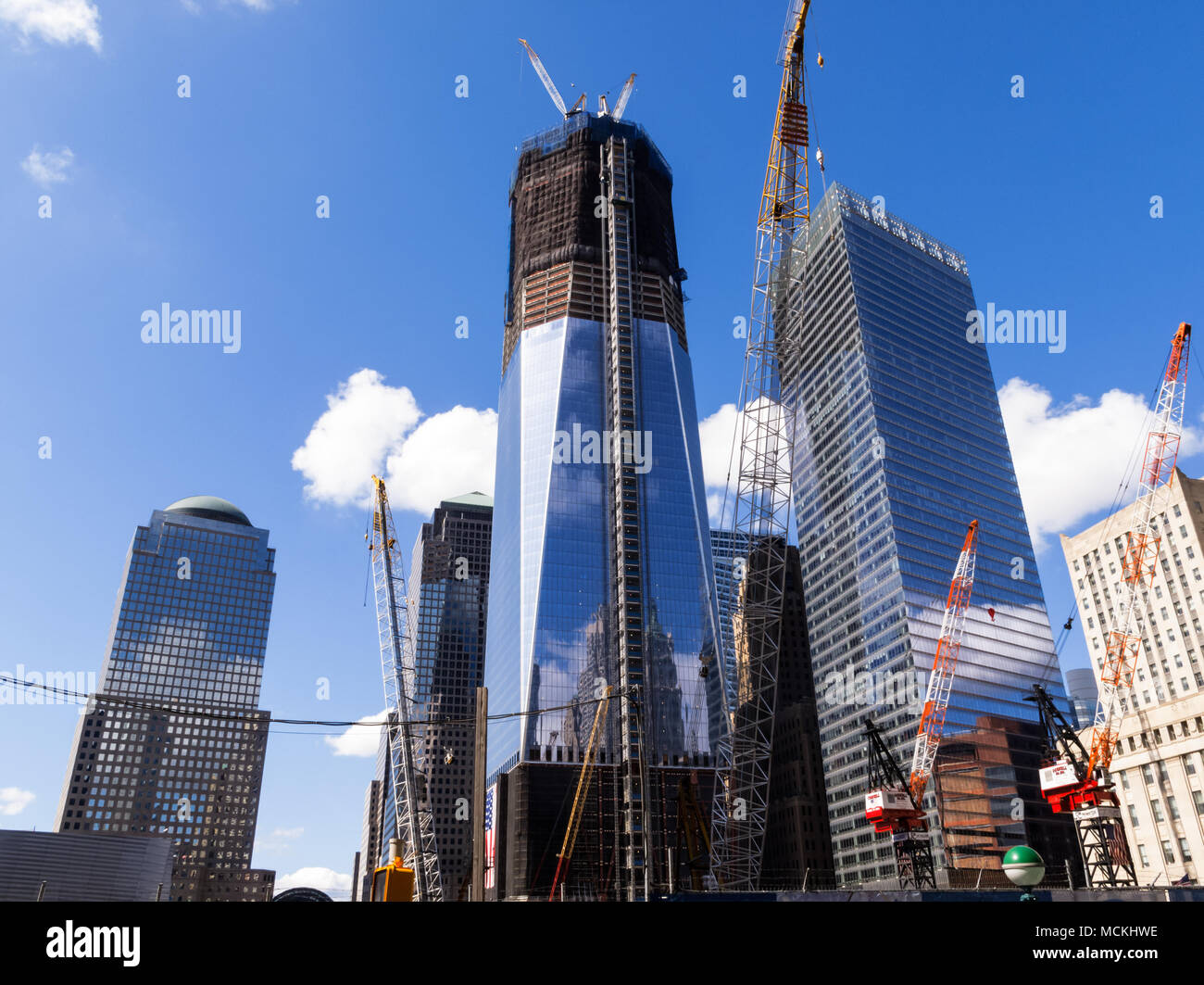 Ein Niedrigwinkelfoto zeigt den laufenden Bau des One World Trade Centers mit Kränen, die aktiv am Bauprozess beteiligt sind, vor dem Hintergrund anderer Wolkenkratzer und eines klaren blauen Himmels. Stockfoto