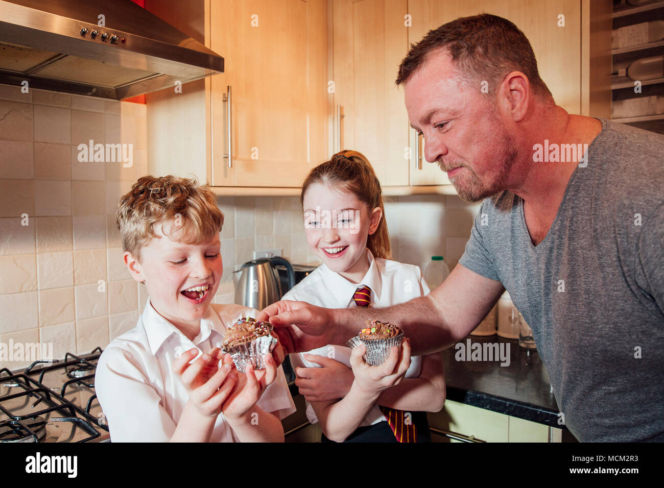 Zwei Kinder sind in der Küche ihres Hauses nach der Schule. Sie essen Cupcakes und der Vater stiehlt einige Tüpfelchen oben von seinen Söhnen cak Stockfoto