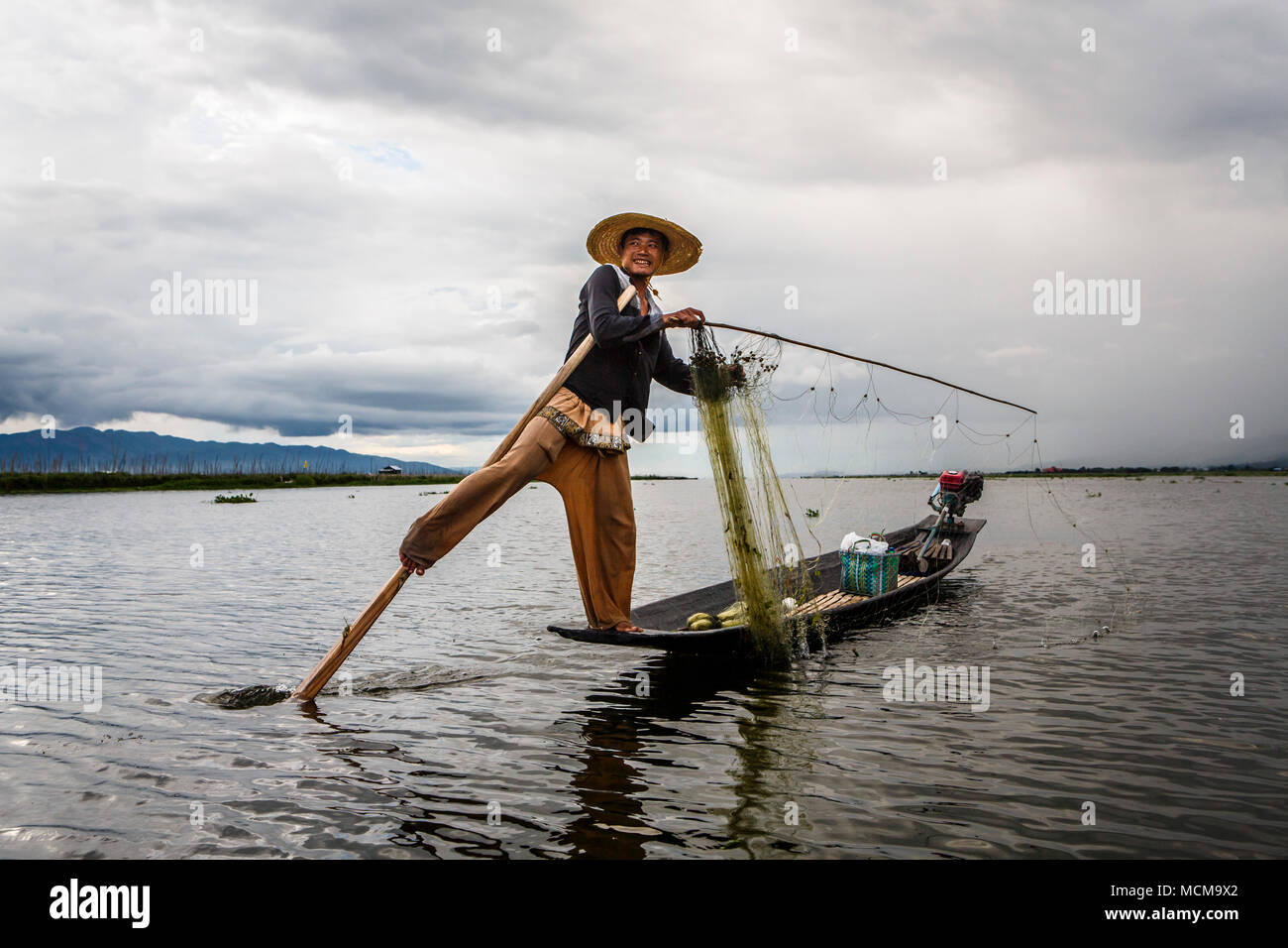 Fischer lächelte Kamera beim Balancieren auf Motorboot mit Angelrute und Net, Shan Staat, Myanmar Stockfoto