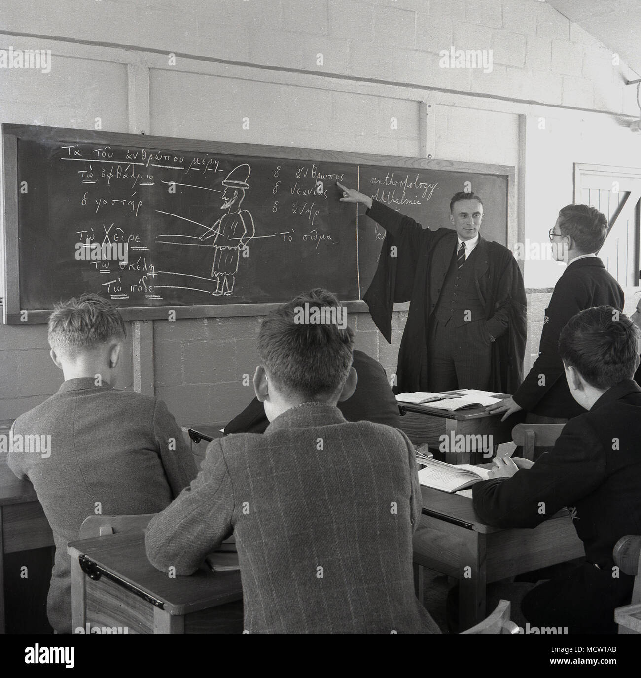 1950er Jahre, historische Bild eines erwachsenen männlichen Lehrer in Hemd und Krawatte und das Tragen eines traditionellen Kleid mit einer großen Tafel zu lehren, eine kleine Gruppe von Schülern am Schreibtisch sitzen, England, UK erziehen oder. Stockfoto