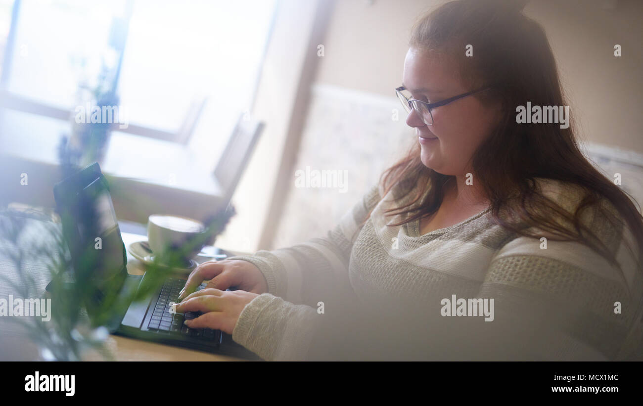 Unscharfer Vordergrund während chubby weiß Frau in ein modernes Cafe, mit hellem Licht sitzt, arbeitet sie an ihrem neuesten Projekt, als sie Arten ihre digitalen Tablet o. Stockfoto