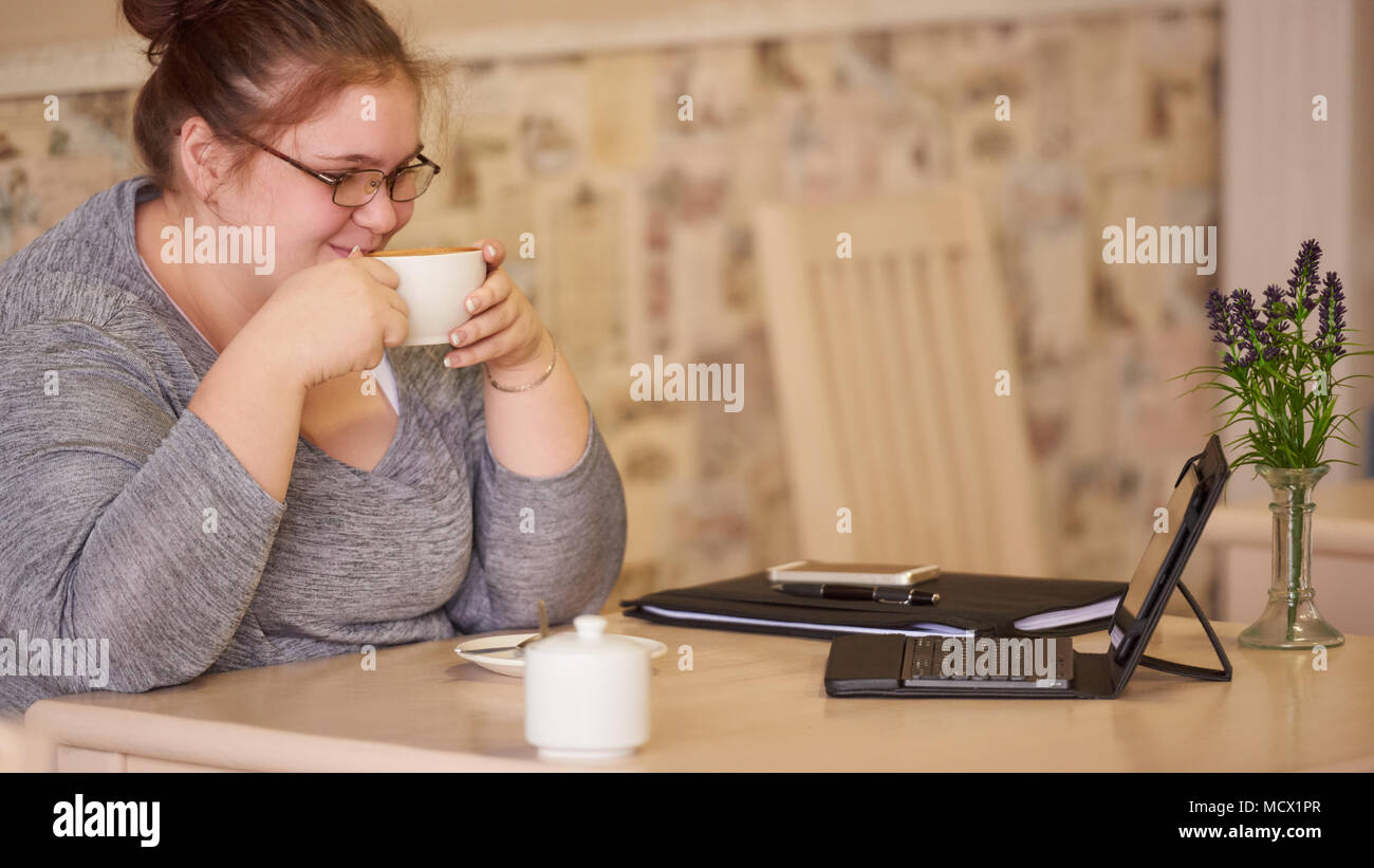 Mäßig adipösen Geschäftsfrau in einem Cafe sitzen mit einer frischen Tasse Kaffee, die Sie hält mit beiden Händen einen Schluck zu nehmen, bevor sie beginnt mit der Arbeit an Ihrem elektronischen Tablet-PC und Handy. Stockfoto