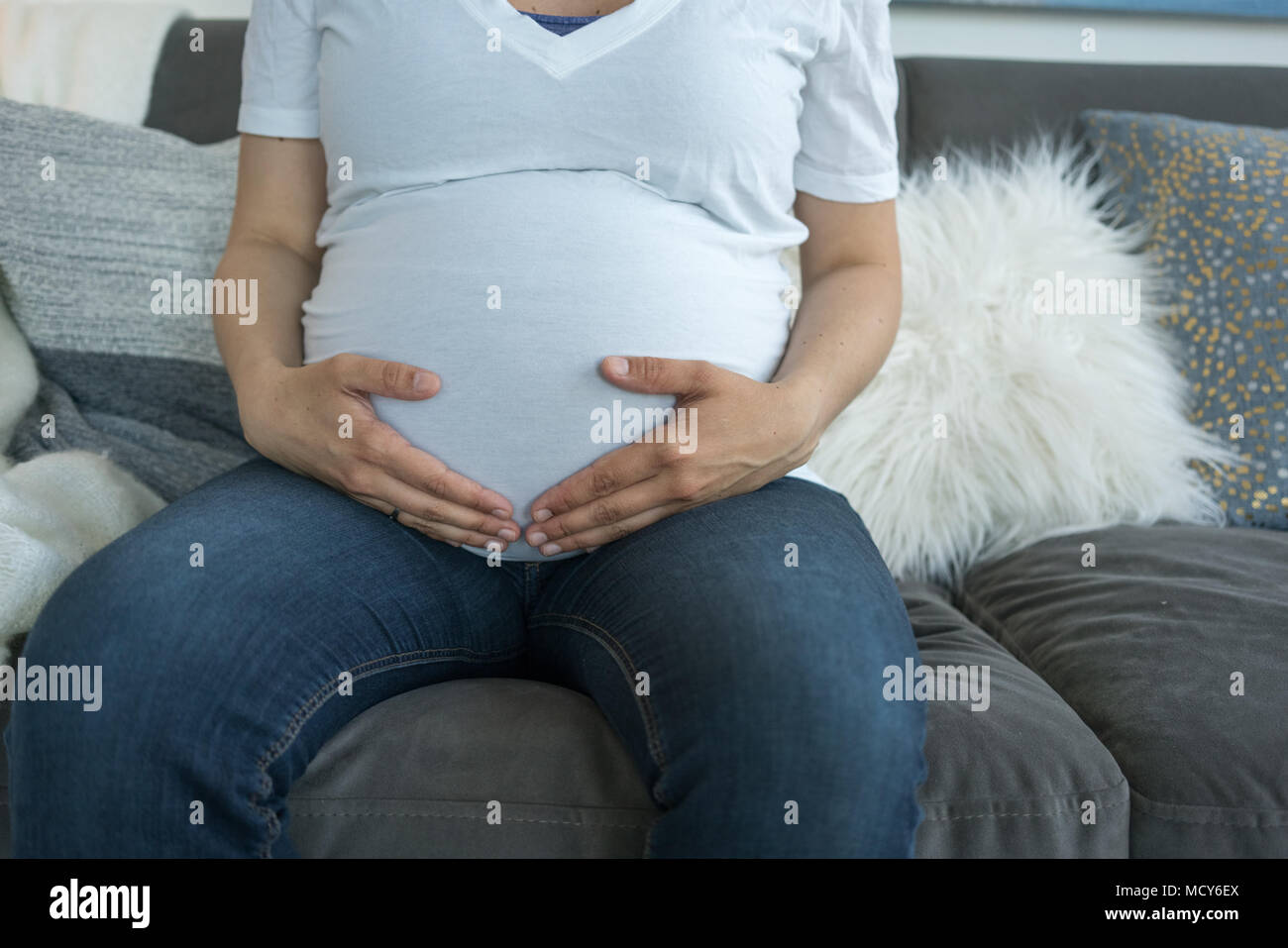 Mittelteil der schwangeren Frau sitzt auf der Couch und ihren Bauch berühren. Stockfoto