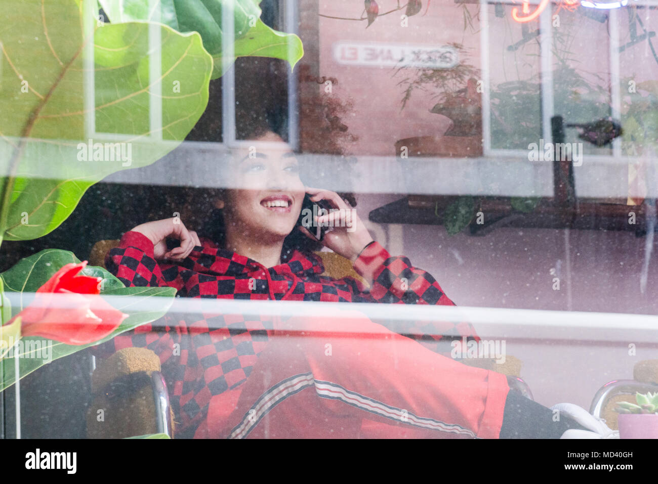 Junge Frau sitzt drinnen, mit Smartphone, durch das Fenster gesehen Stockfoto