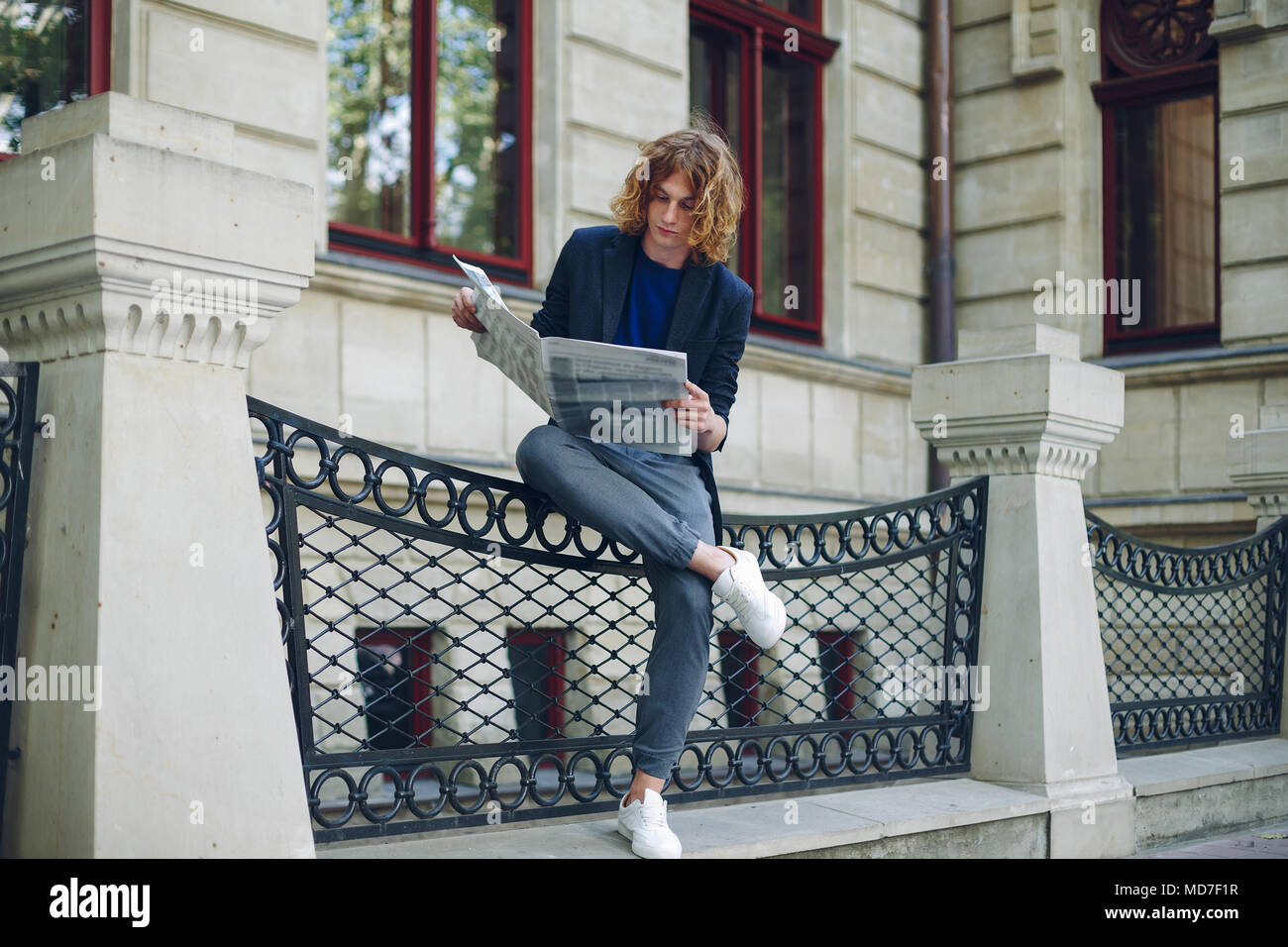 Junge rötlich, rothaarige attraktiver Mann mit lockigem Haar, eine Zeitung lesen sitzen Bein über Bein in der Nähe einer städtischen alten Gebäude. Jugend in Aktion y Stockfoto