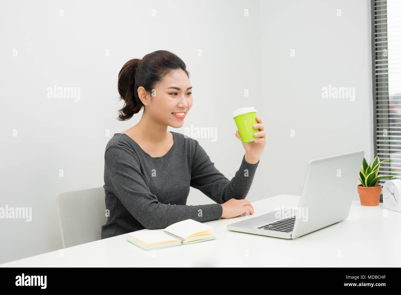 Junge asiatische Frau Unternehmer an einem Tisch sitzen in Ihrem Büro zu Hause trinken einen Kaffee zum Mitnehmen und online arbeiten mit einem Laptop und einer Tablette Stockfoto