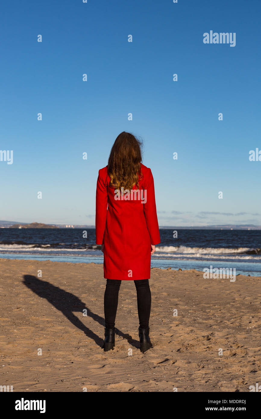 Junge Frau am Strand, trägt eine rote Jacke, mit ihr zurück gedreht, mit Meerblick. Hochformat. Stockfoto