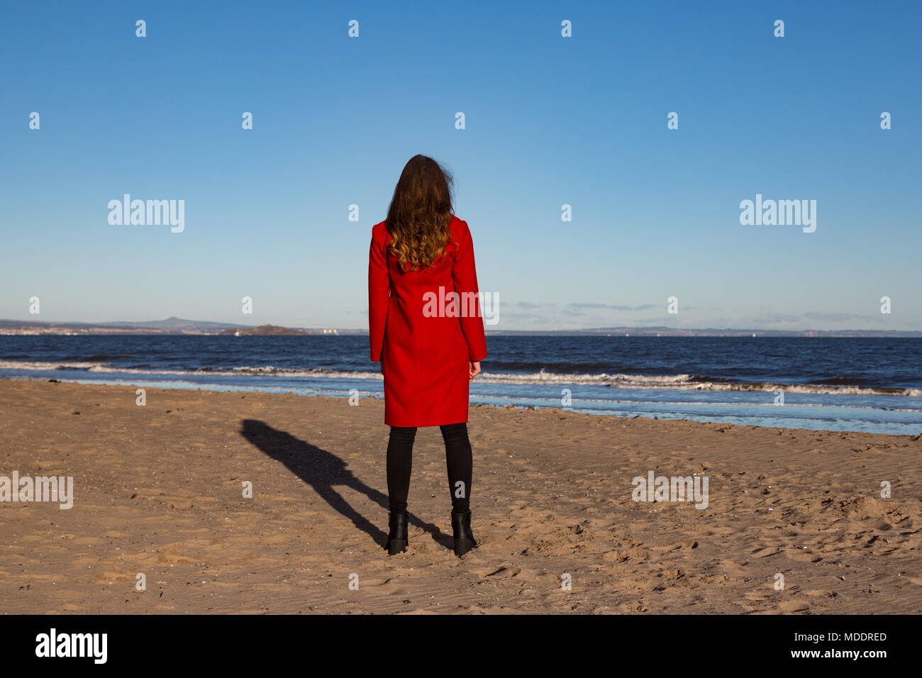 Junge Frau am Strand, trägt eine rote Jacke, mit ihr zurück gedreht, mit Meerblick. Querformat. Stockfoto