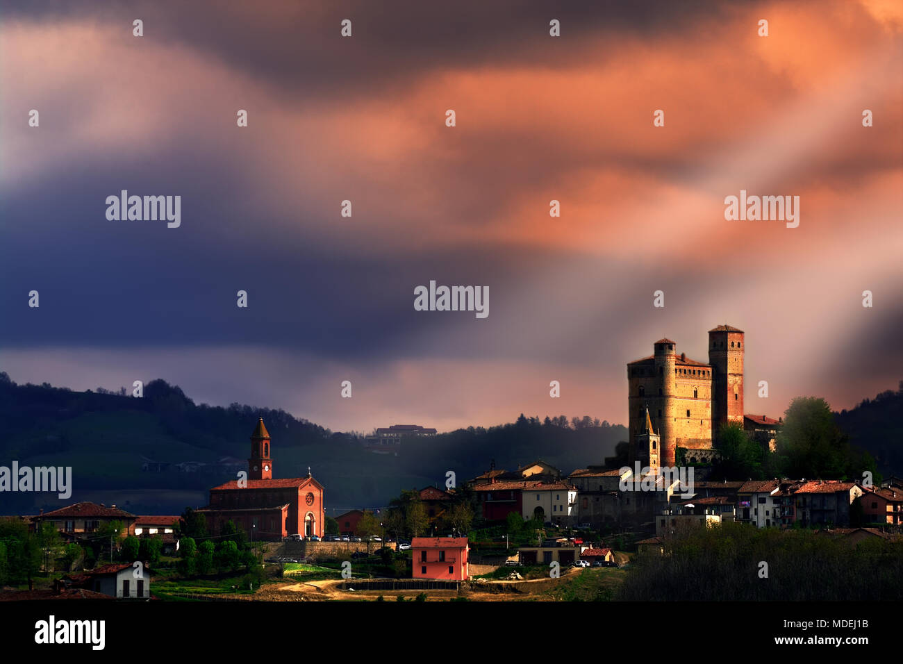 Sonne strahlen leuchtet auf, das Schloss und die Kirche von Serralunga d'Alba unter einem stürmischen Himmel. Langhe, Piemont, Italien. UNESCO-Weltkulturerbe. Stockfoto