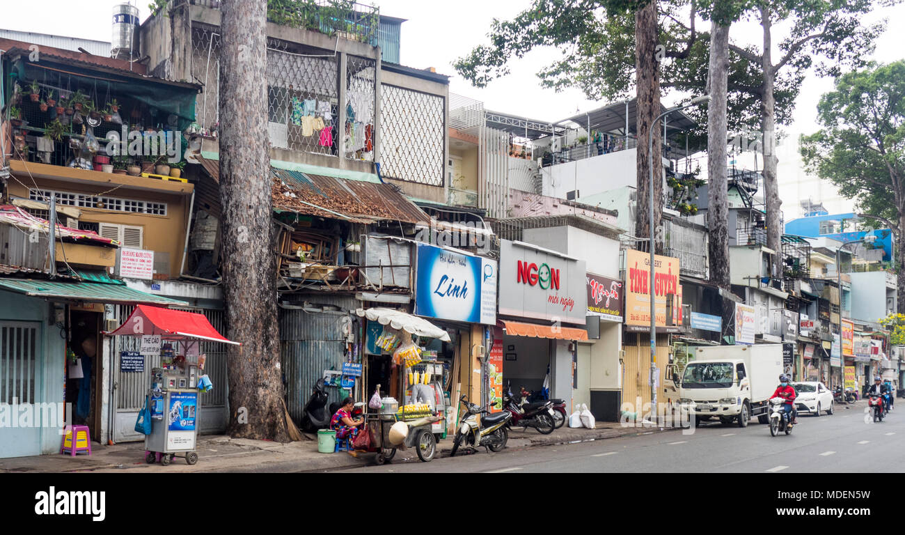 Straßenbild aus einer Reihe von Geschäftshäusern und Street Food Anbieter auf einer von Bäumen gesäumten Straße in Ho Chi Minh City, Vietnam. Stockfoto