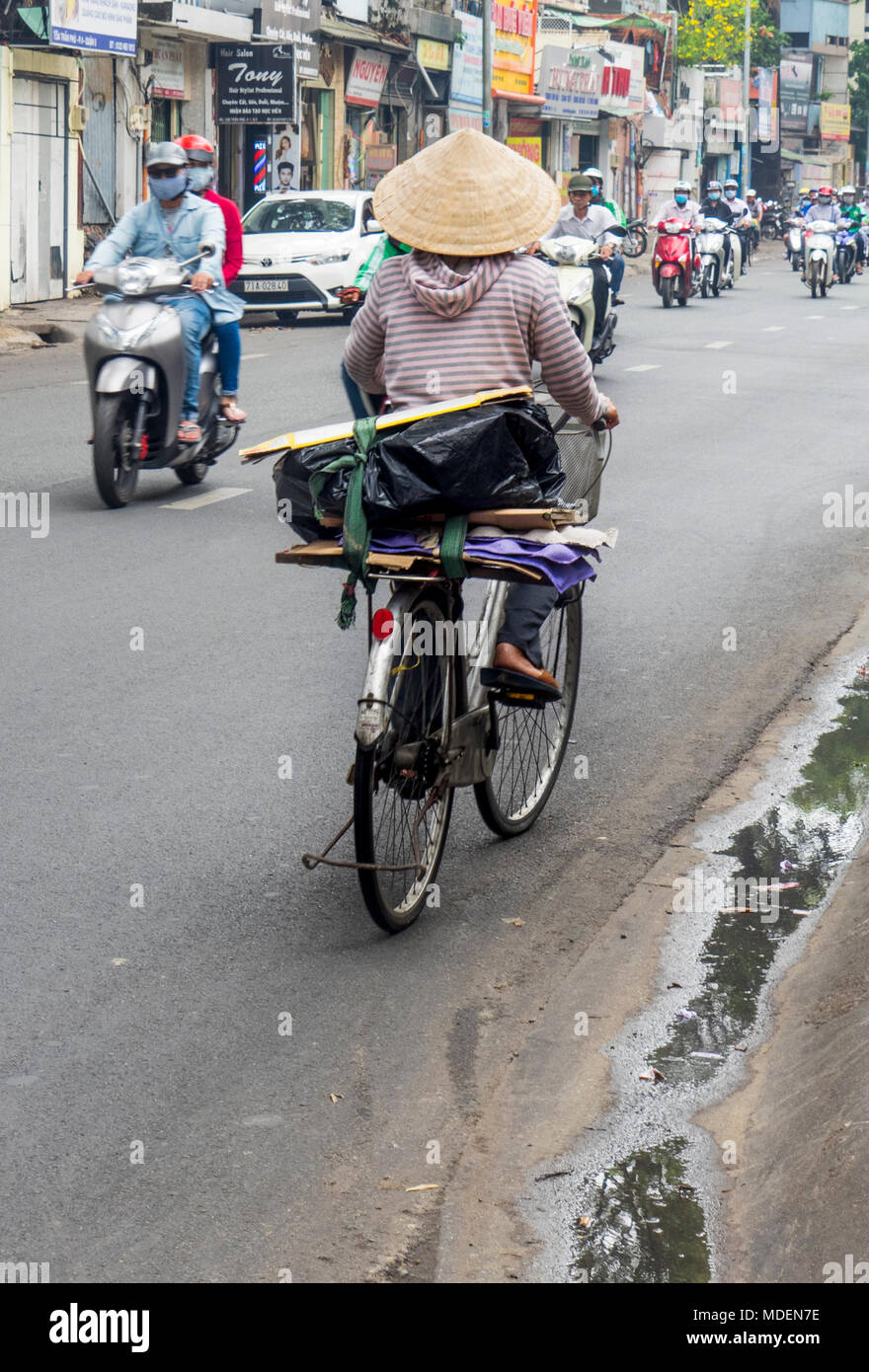 Eine vietnamesische Frau Radfahrer tragen eine kegelförmige Hut reiten ihr Fahrrad auf einer Straße in Ho Chi Minh City, Vietnam. Stockfoto