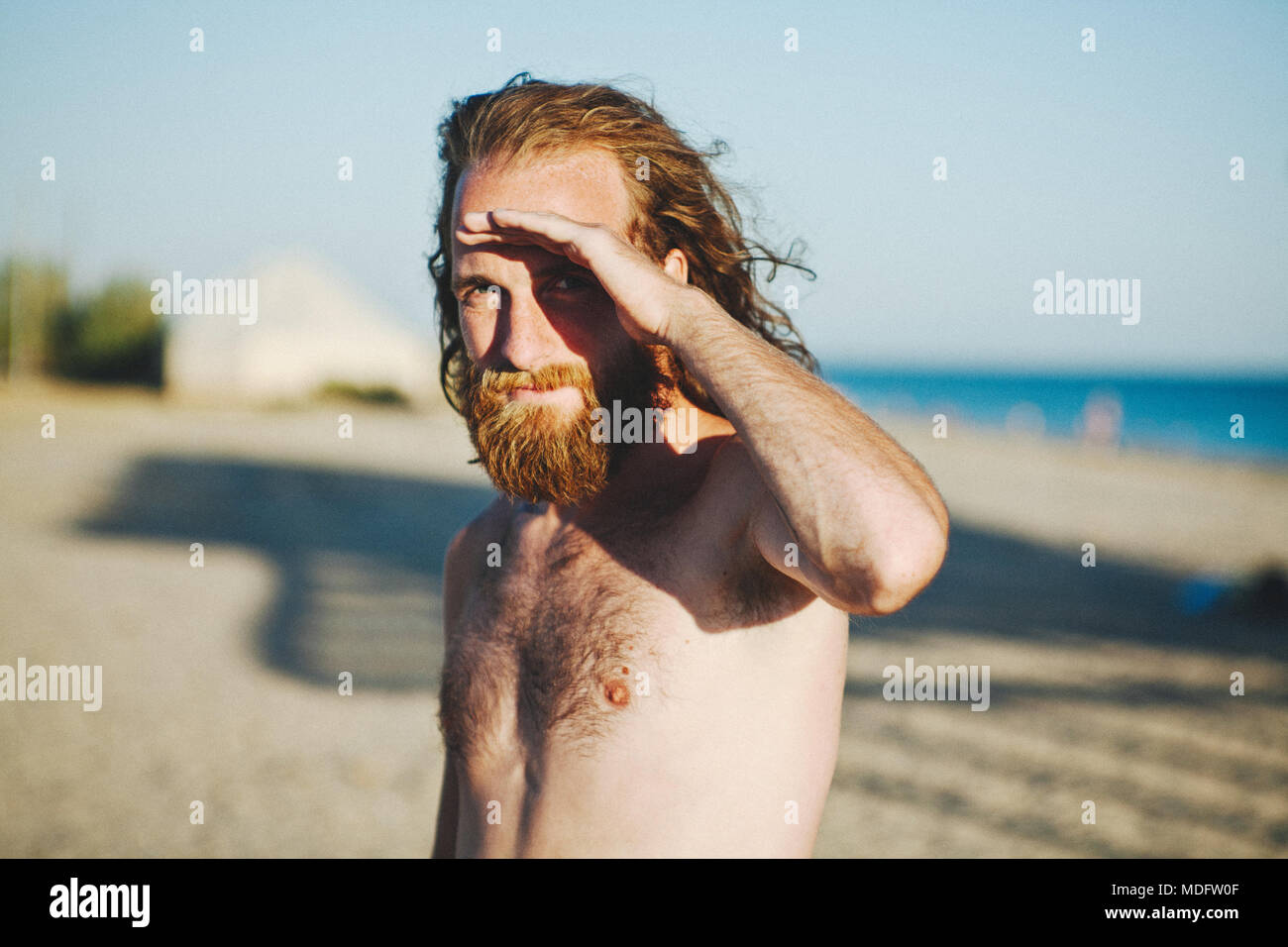 Mann mit langen Haaren und Bart steht auf Strand Abschirmung seine Augen vor der Sonne Stockfoto