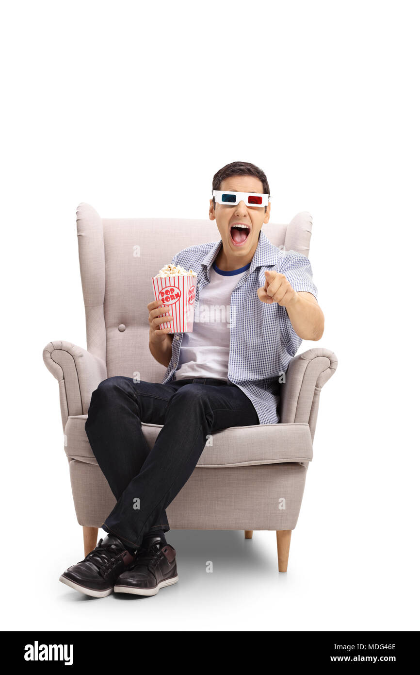 Junger Mann mit 3D-Brille und Popcorn in einem Sessel lachen und zeigen auf die Kamera auf weißem Hintergrund sitzt Stockfoto