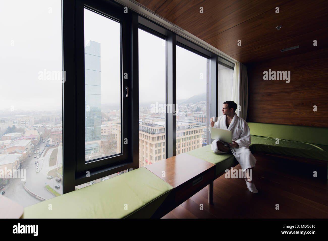 Unternehmer zu Hause oder in der Reise. Seitenansicht Portrait von hübscher junger Geschäftsmann sitzt auf dem Bett und mit Laptop. Panoramafenster mit schönen Sonnenaufgang Stadt Landschaft im Hintergrund Stockfoto