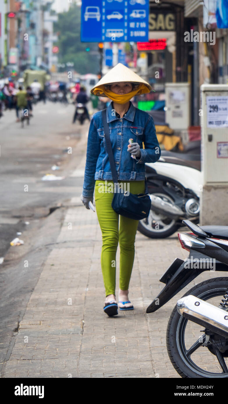 Eine vietnamesische Frau trug eine kegelförmige Hut, Jeans Jacke und grüne Hose zu Fuß auf dem Gehweg auf einer Straße in Ho Chi Minh City, Vietnam. Stockfoto