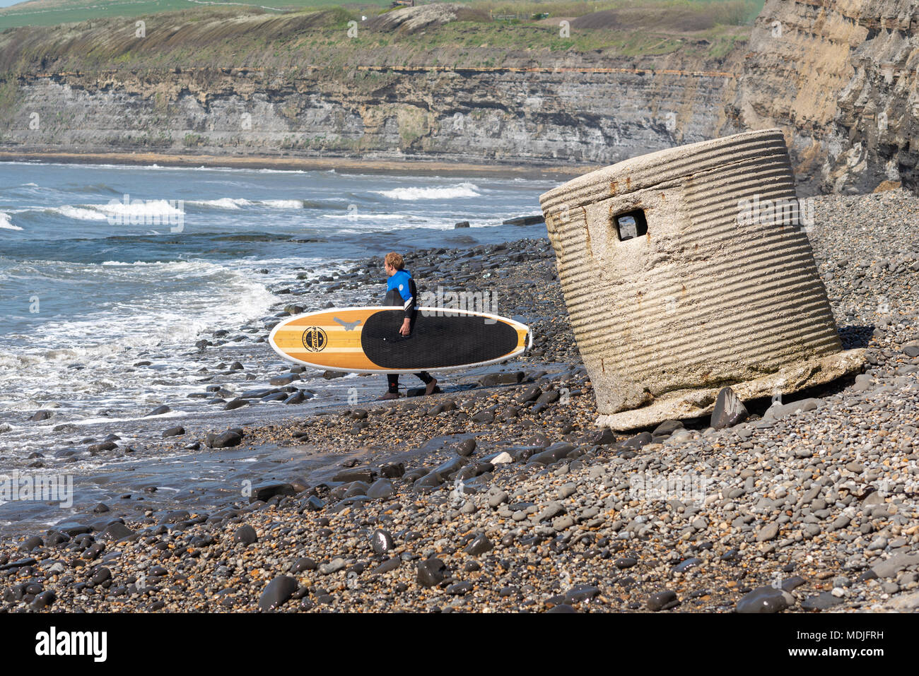 Ein Surfer geht raus auf das Meer mit seinem Surfboard unter dem Arm vorbei an der Ruine einer WWII Pillenschachtel lookout Post am Strand bei Kimmeridge Bay, Dorset. Stockfoto