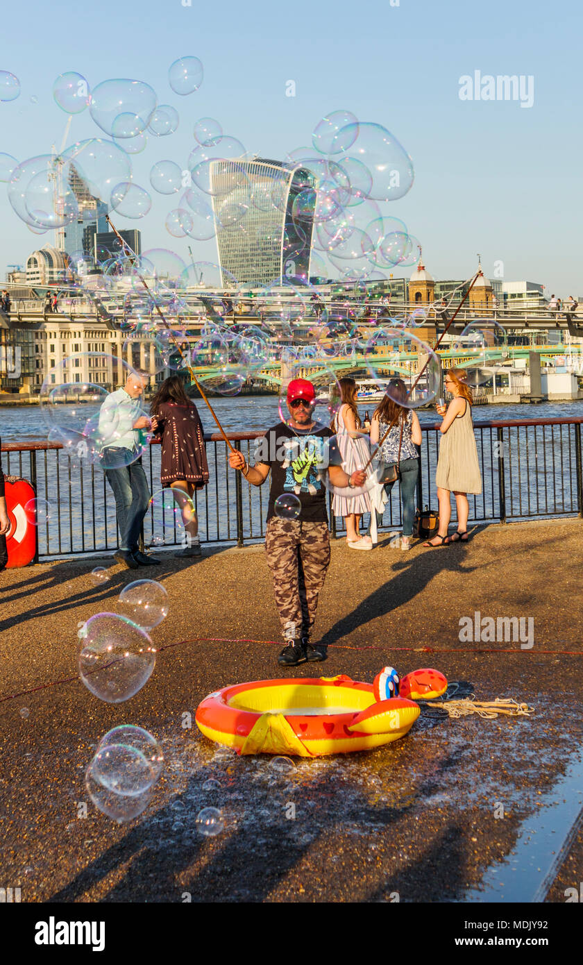 London, UK, 19. April 2018. Ein Entertainer am Südufer der Ufer der Themse in Bankside bläst Seifenblasen Kinder gegen eine Skyline Hintergrund von Ikonischen moderne Wolkenkratzer Gebäude in der Stadt London zu unterhalten. Die Sonnige schönem Wetter auf den wärmsten April Tag seit Jahrzehnten brachten gutmütige Massen, die Sonne zu genießen. Credit: Graham Prentice/Alamy Leben Nachrichten. Stockfoto