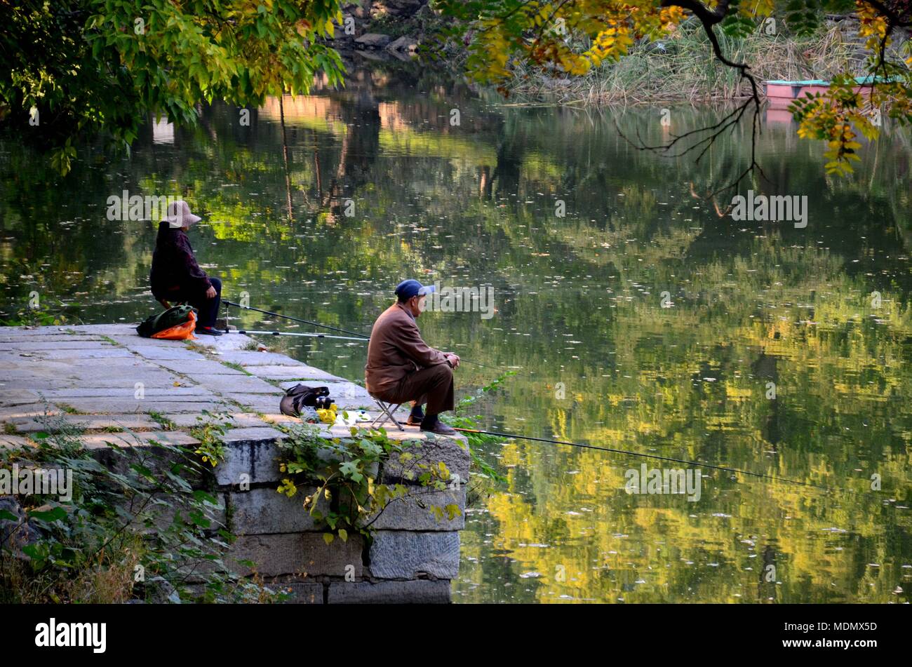 Paar Hüte tragen sitzen ruhig und separat auf Hockern an Steg und Fisch an einem See außerhalb von Peking, China Stockfoto