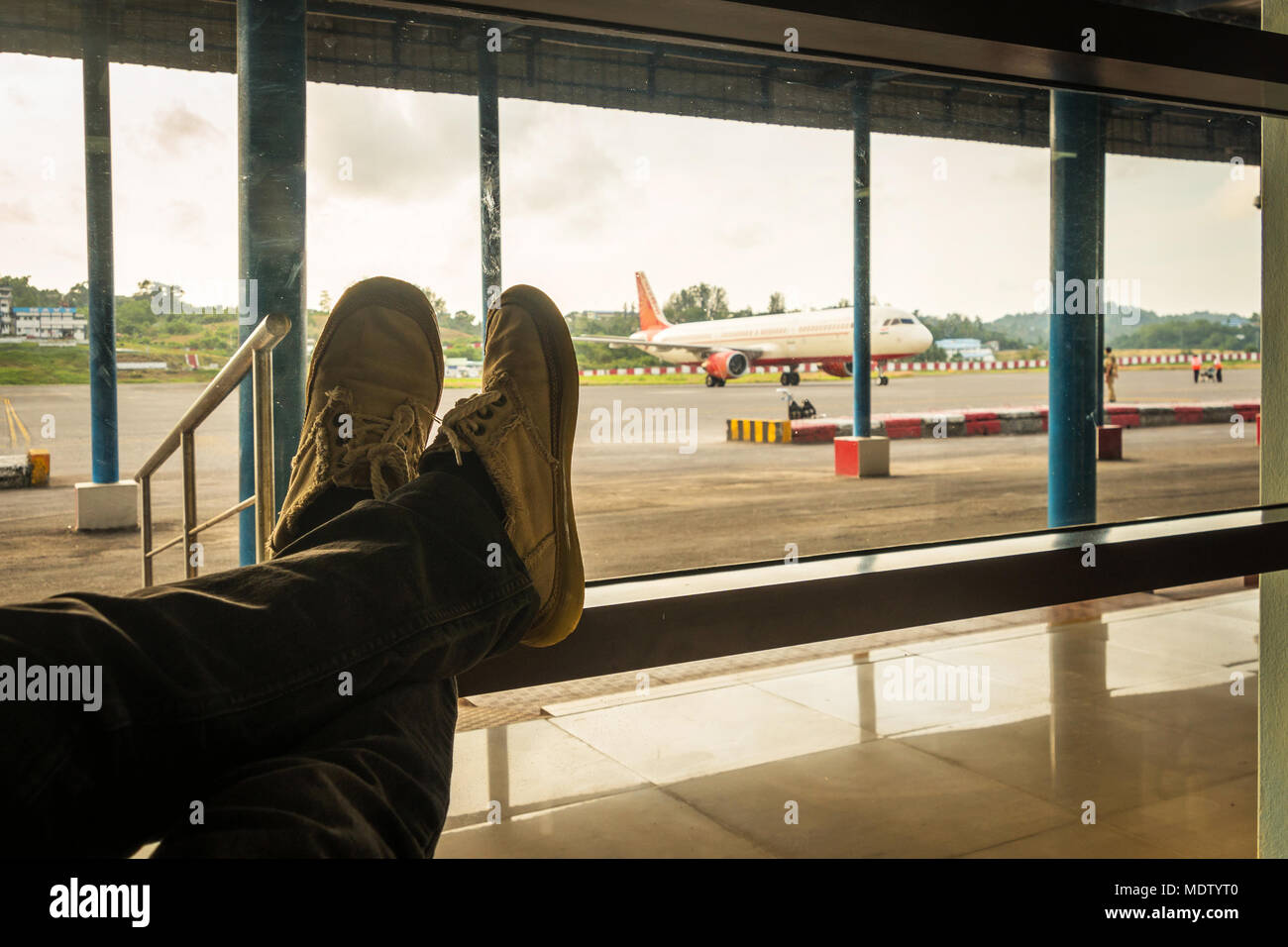 Männliche warten auf Ihren Flug. Der Passagier sitzt am Flughafen mit seinem Bein vor dem Hintergrund von Flugzeugen auf dem Rollfeld gefaltet. Stockfoto