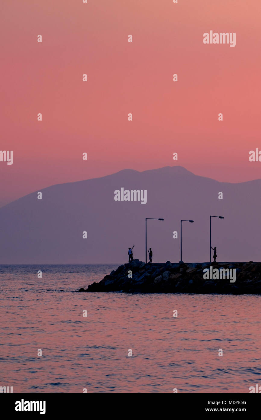 Menschen Silhouetten unter selfies auf einen Wellenbrecher bei Sonnenuntergang in warmen lila Licht. Die Berge im Hintergrund. Stockfoto