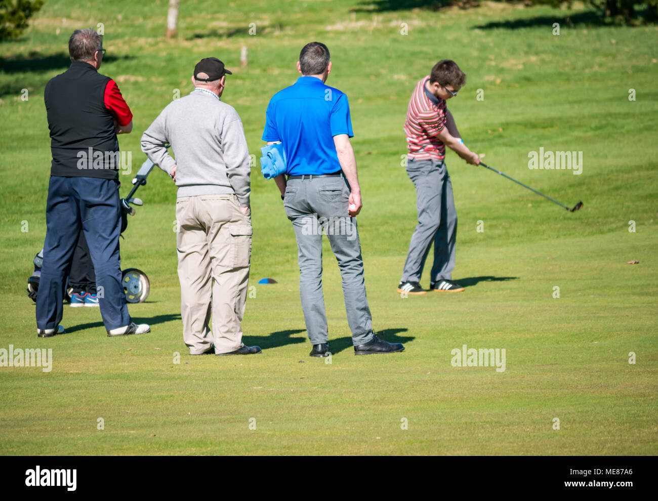 West Linton, Scottish Borders, Schottland, Vereinigtes Königreich, 21. April 2018. Frühlingssonne in der Landschaft, mit einer Gruppe von männlichen Golfspieler auf einer Fahrrinne an der West Linton Golfplatz. Ein junger Mann ist das Schwingen einer Golf Club, von drei Männern beobachtet Stockfoto