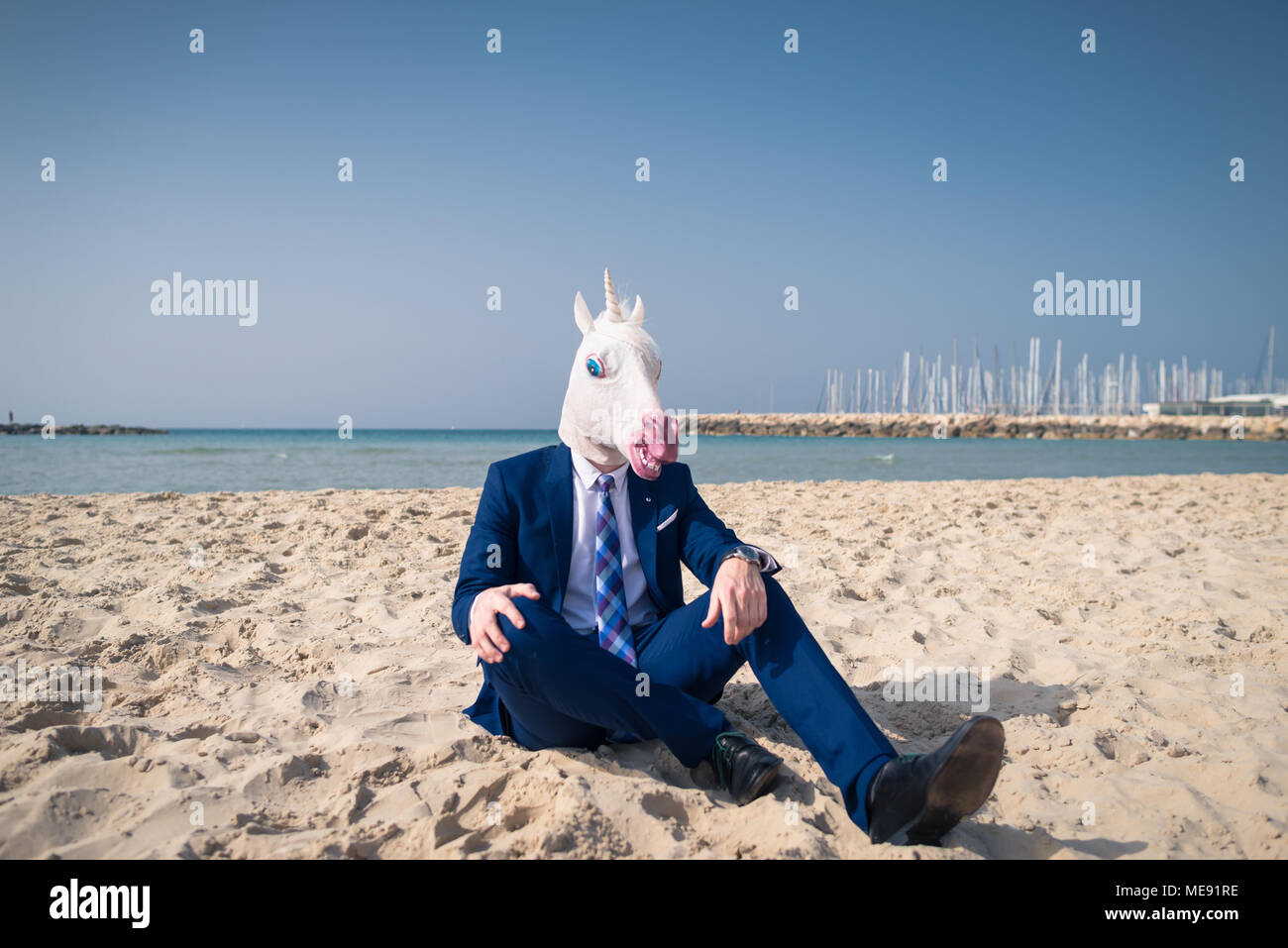 Junger Mann in lustige Maske sitzt auf dem Sand und genießt Urlaub. Ungewöhnliche Kerl in stilvollen Anzug entspannt am Strand. Einhorn auf Hintergrund von Himmel und Meer Stockfoto