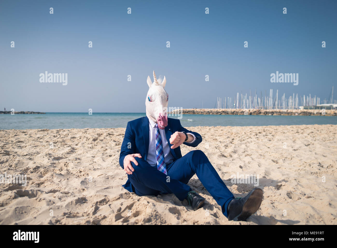 Ernster Mann in komischen Maske sitzt auf dem Sand und genießt sonnigen Tag. Ungewöhnliche Kerl in Anzug auf der Suche nach Armbanduhr. Seltsame person Relaxen am Strand Stockfoto
