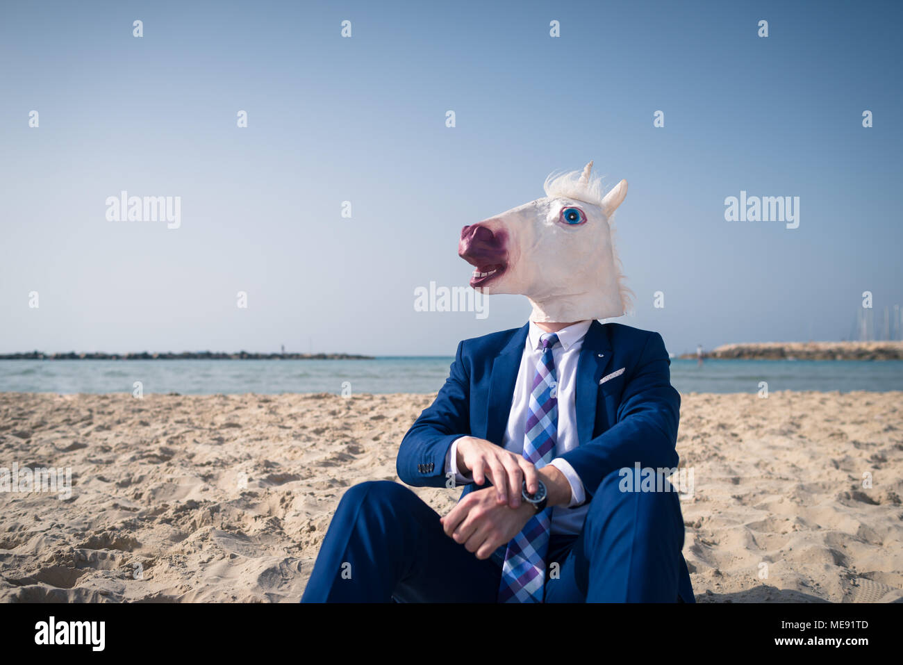 Stilvolle Mann in lustige Maske sitzt auf dem Sand und genießt sonnigen Tag. Ungewöhnliche Mann im eleganten Anzug entspannt am Strand. Einhorn auf Hintergrund von Himmel und Meer Stockfoto